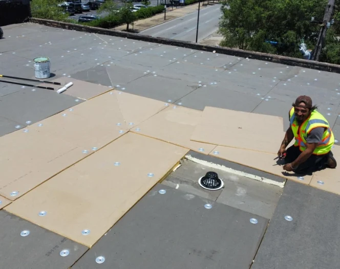 A smiling worker installs roofing insulation boards on a commercial roof, with tools and a drain visible.